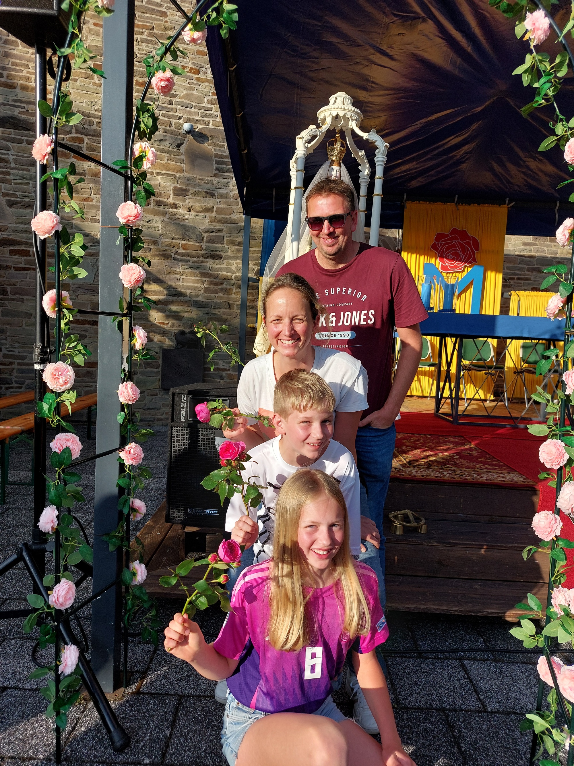 Familie Müller steht hintereinander unter einem Rosenbogen vor der Rosa Mystica. Jeder hält eine Rose in der rechten Hand.